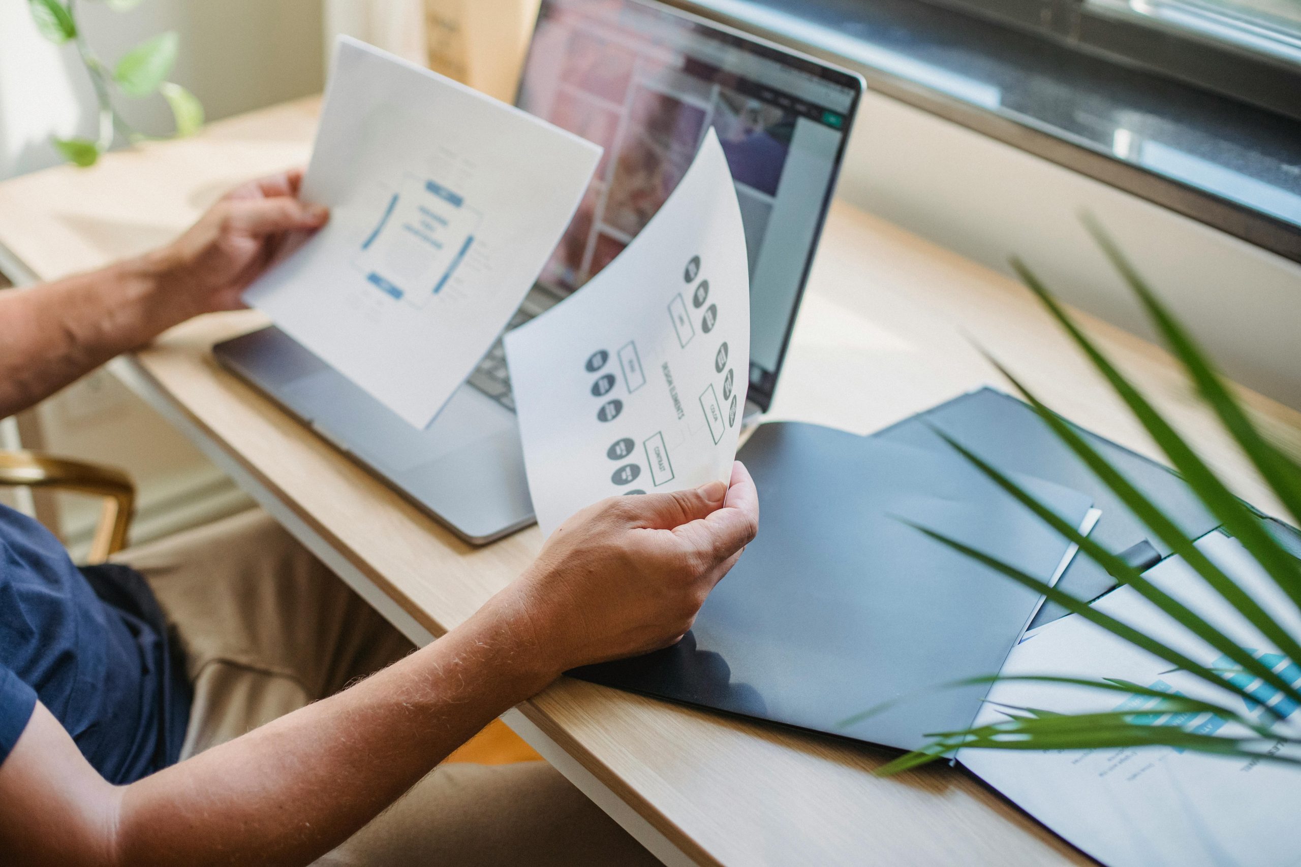 A small business owner sits at a desk, considering the pros and cons of IT outsourcing. A laptop and paperwork are spread out in front of them. Outside the window, a remote worker can be seen on a computer